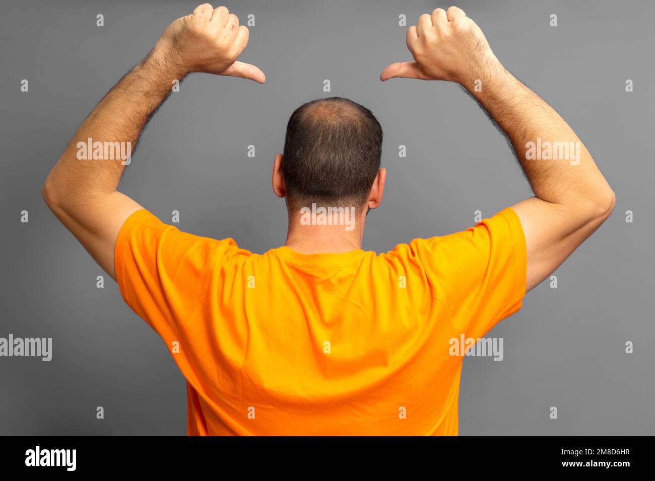 Proud man in orange t-shirt with his back turned pointing with his thumbs at his receding bald spot, isolated on gray background. Stock Photo