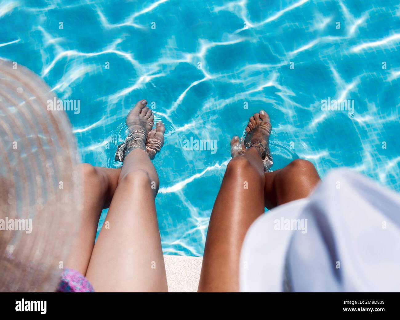 rear view of two girls of different ethnicity are sitting with their feet in the water at the edge of a pool and only their legs are visible. concept Stock Photo