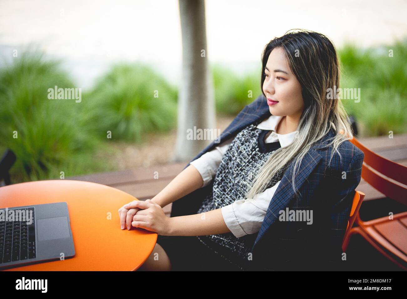 Young Woman Sitting Outdoors at an Orange Metal Table with Laptop Dressed for Autumn Weather Stock Photo