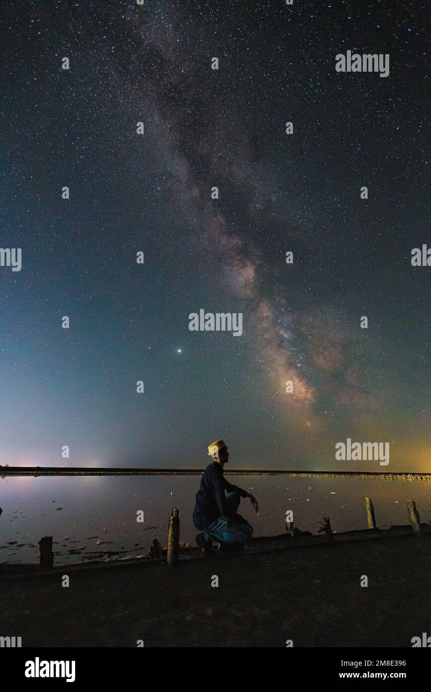 Man in the hat sit and look up at the sky. Bright night landscape with Milky Way galaxy stripe and Jupiter over the salt lake in Henichesk, Ukraine Stock Photo