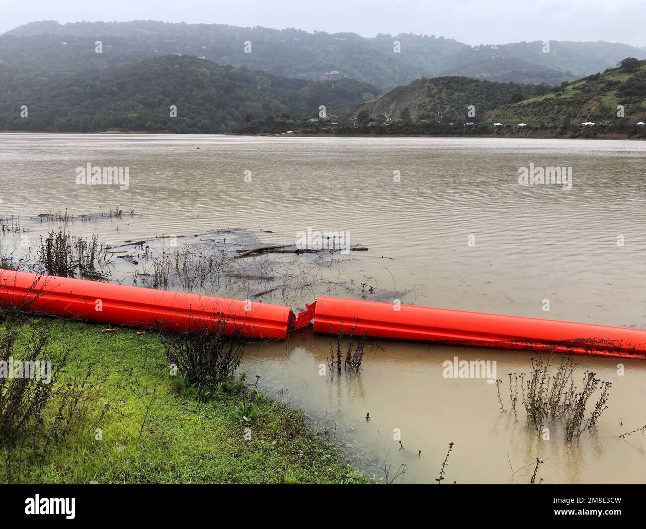 Orange floating debris boom, barrier on the green grass and in the water of a full reservoir during the rainy season Stock Photo
