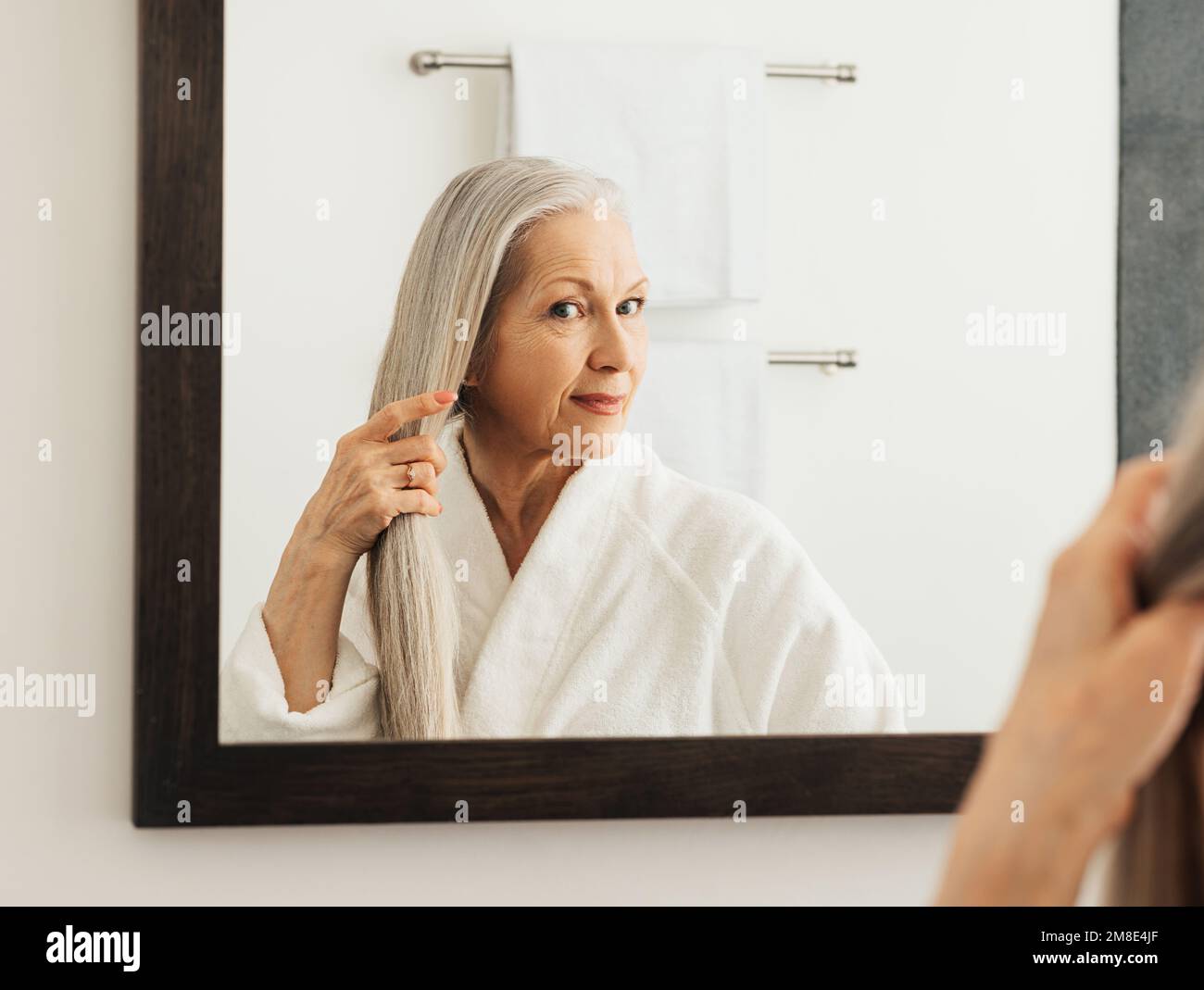 Aged woman with long gray hair looking at a mirror in the bathroom ...