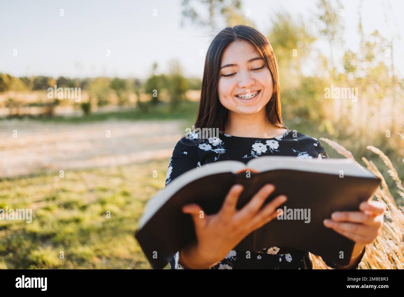 Young religious christian girl holding and reading her bible, outside in the field at sunset. Communion with God. Stock Photo