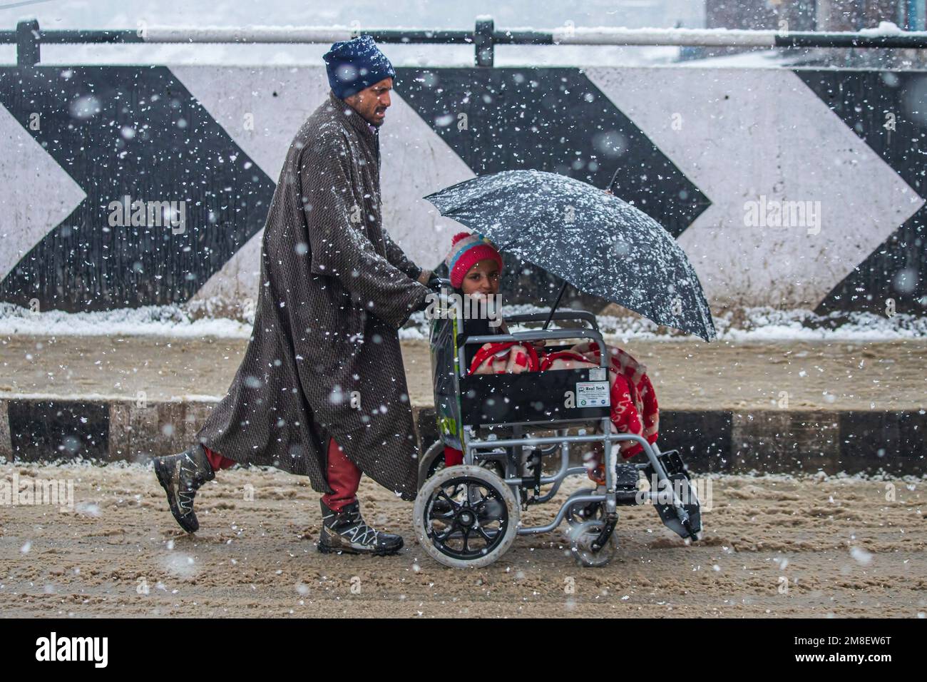 A father carrying his allying son on a wheel chair as he walks through a street amid ongoing heavy snowfall on the outskirts of Srinagar. At least two labourers killed on Thursday after an avalanche hits high-altitude area in central Kashmir's Sonamarg. Most parts of Kashmir received fresh snowfall that leading to the closure of Kashmir capital's main highway that connects disputed region with the rest of India. Meanwhile airport authorities suspended all flight operations till further notice due to continuous snowfall and low visibility. (Photo by Faisal Bashir/SOPA Images/Sipa USA) Stock Photo