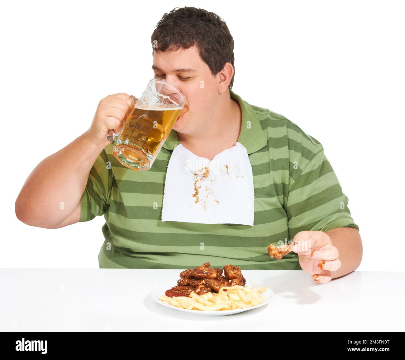 Feasting on fat. An obese young man wearing a bib and drinking a beer with a plate of chicken wings and fries in front of him. Stock Photo