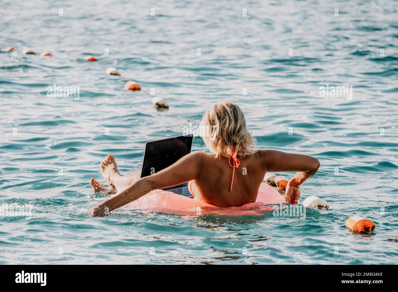 Woman works on laptop in sea. Freelancer, young blond woman in sunglases floating on an inflatable big pink donut with a laptop in the sea at sunset Stock Photo