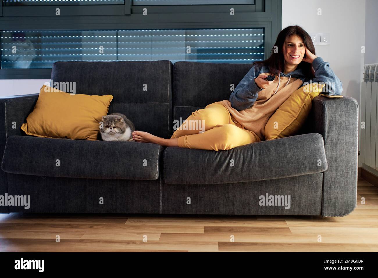 Relaxed woman smiling while watching the tv with her cat on the sofa. Stock Photo