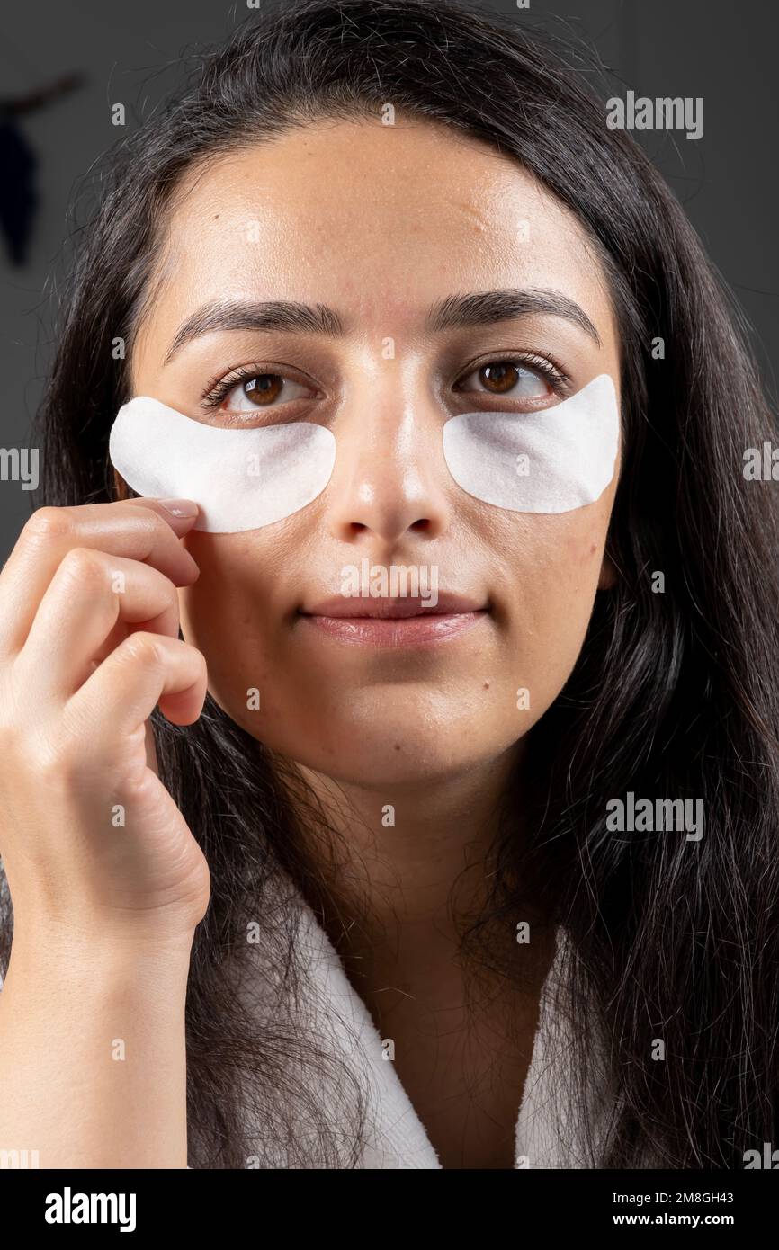 Under eye patches, close up caucasian millennial brunette woman applying under eye patches. Looking mirror using anti aging cosmetic skin care routine Stock Photo