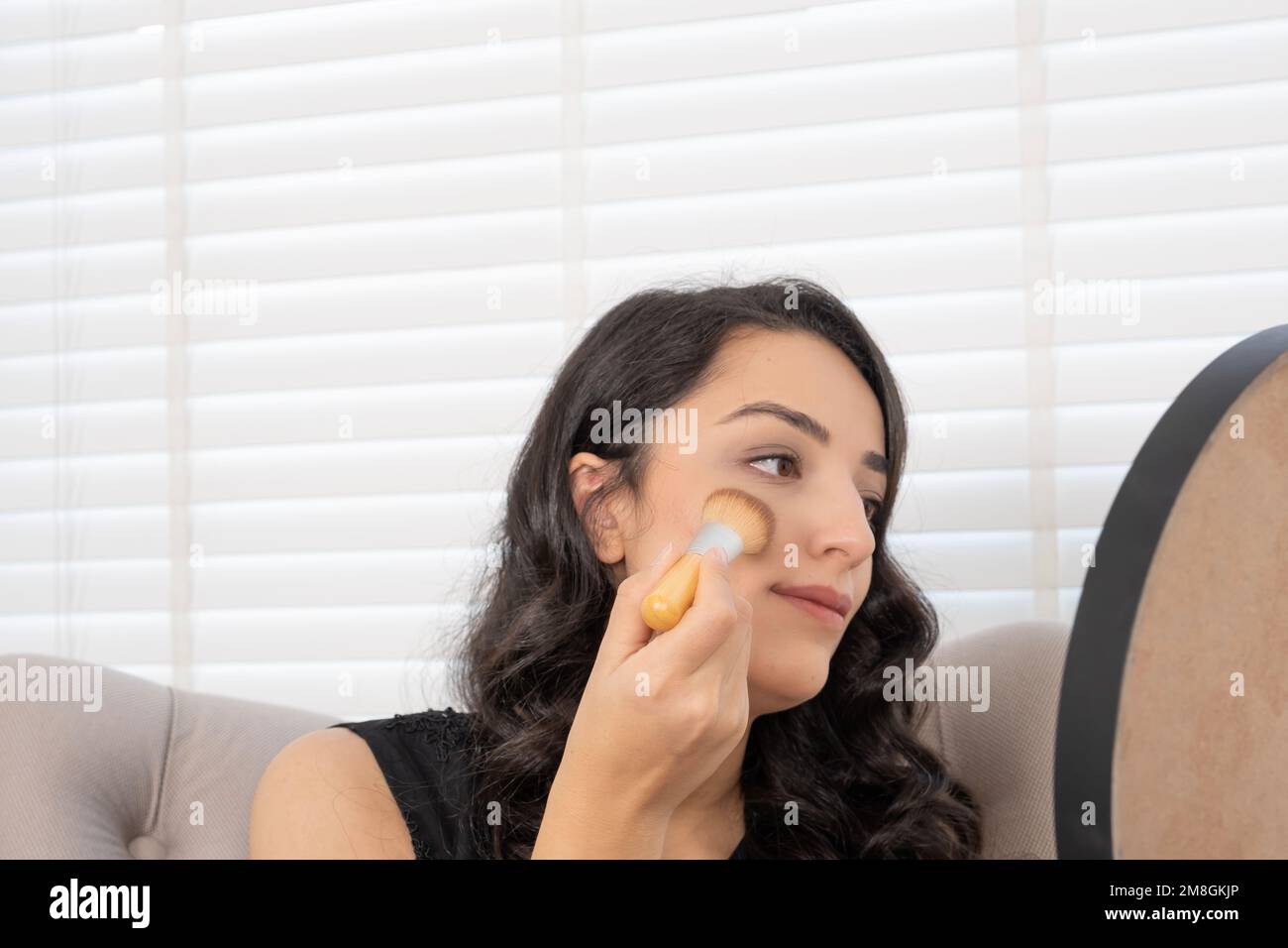Applying blush, brunette millennial woman. Beige foundation powder on cheeks. Using make up brush. Sitting armchair at home. Putting make up concept. Stock Photo