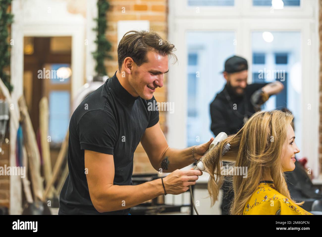 joyful hairdresser drying and combing blond woman's hair, another barber working in the background. High quality photo Stock Photo