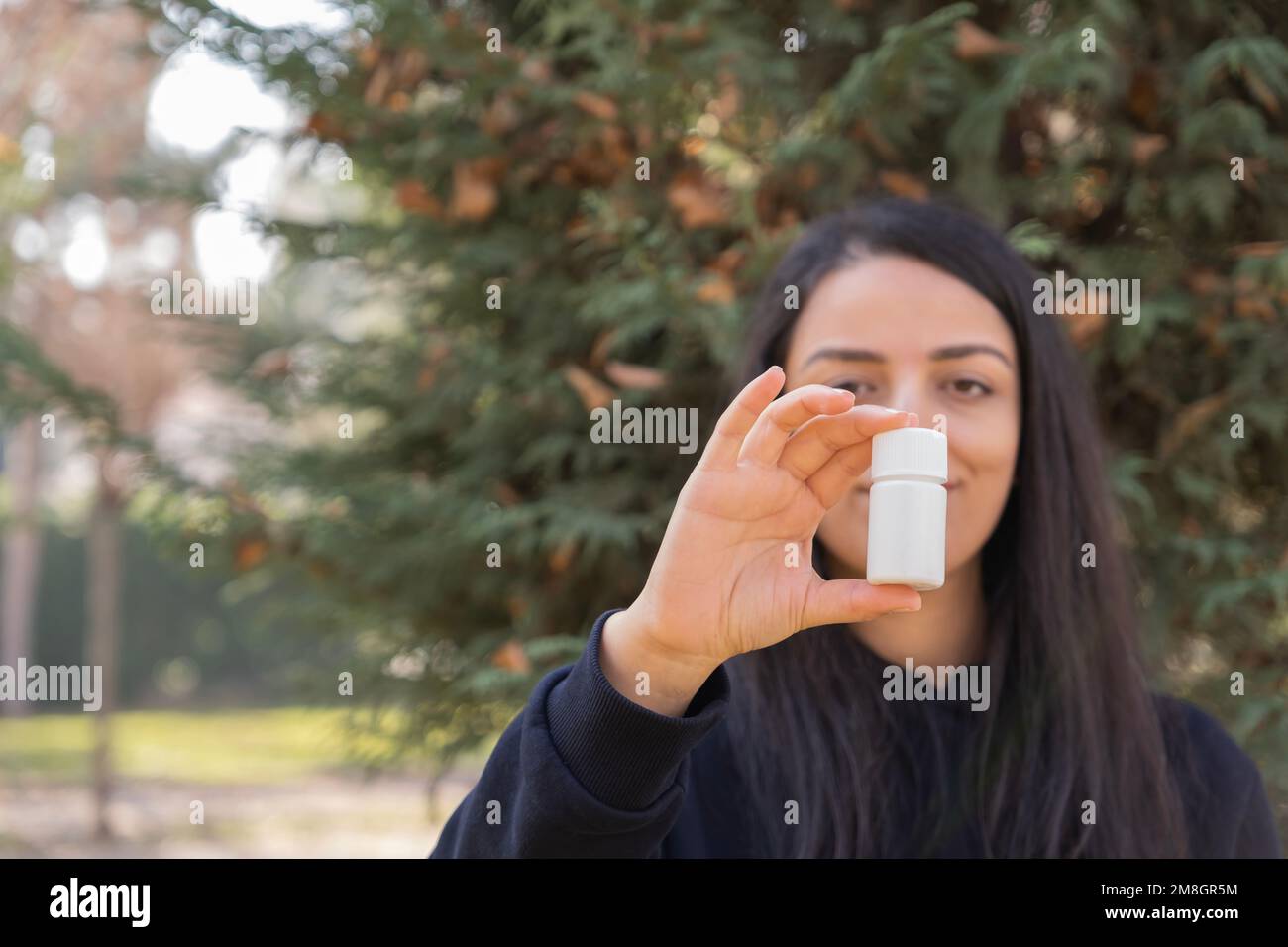 Woman holding white pill bottle for mock up. Beautiful brunette caucasian girl standing before autumn nature background. Copy space medicine concept. Stock Photo