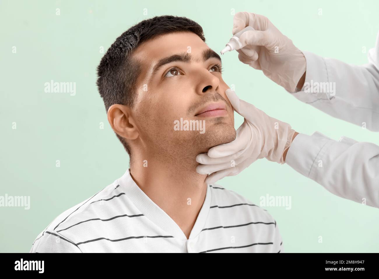 Ophthalmologist putting drops in young man's eye on green background Stock Photo