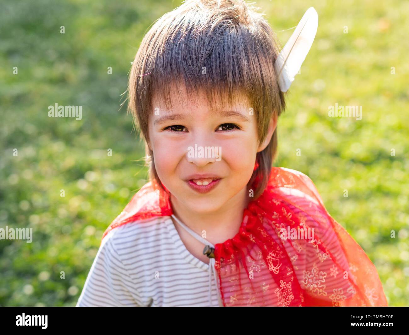 Portrait of smiling boy playing American Indian. Kid with white bird feather and red cloak. Costume role play. Outdoor leisure activity. Stock Photo