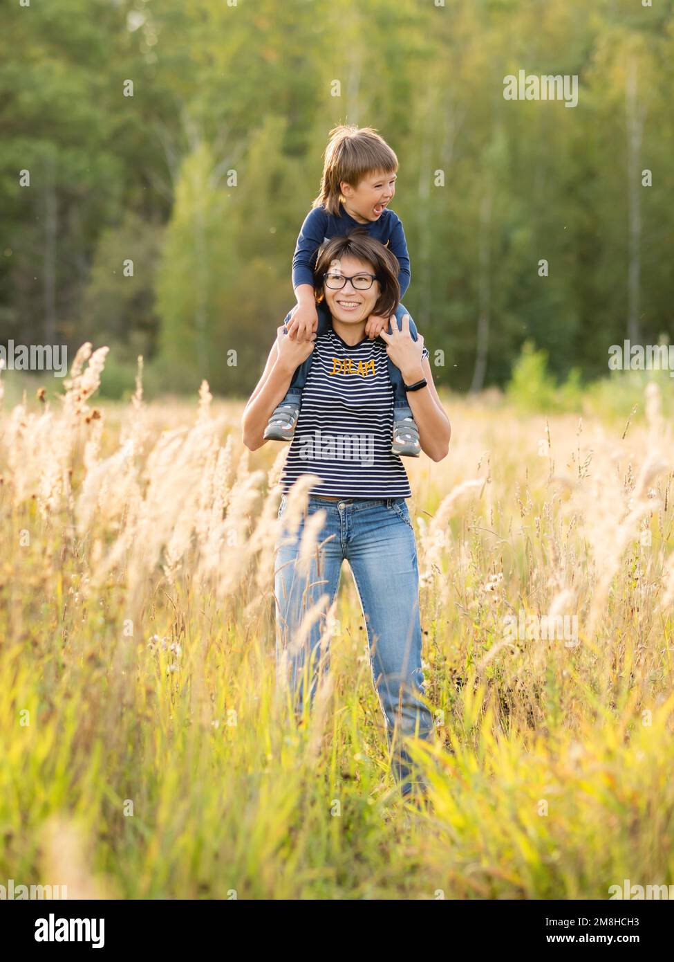 Mother holds her son on shoulders. Happy kid smiles. Mom and boy on field at golden sunset hour. Family life. Autumn season. Stock Photo