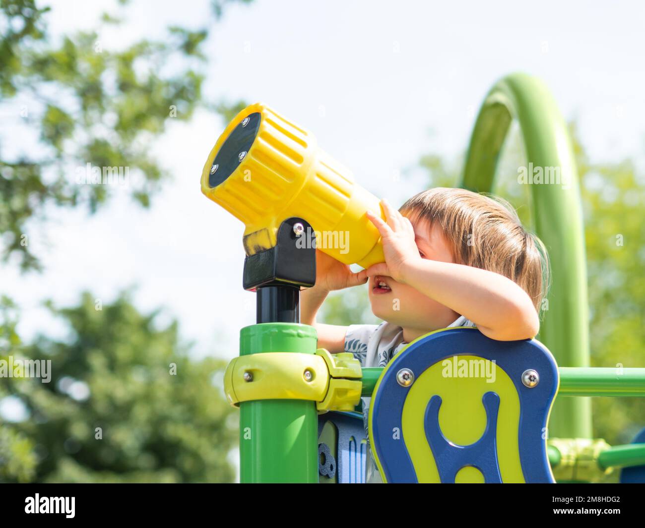 Little kid looks in toy telescope on outdoor playground. Leisure activity for curious children. Summer fun. Stock Photo