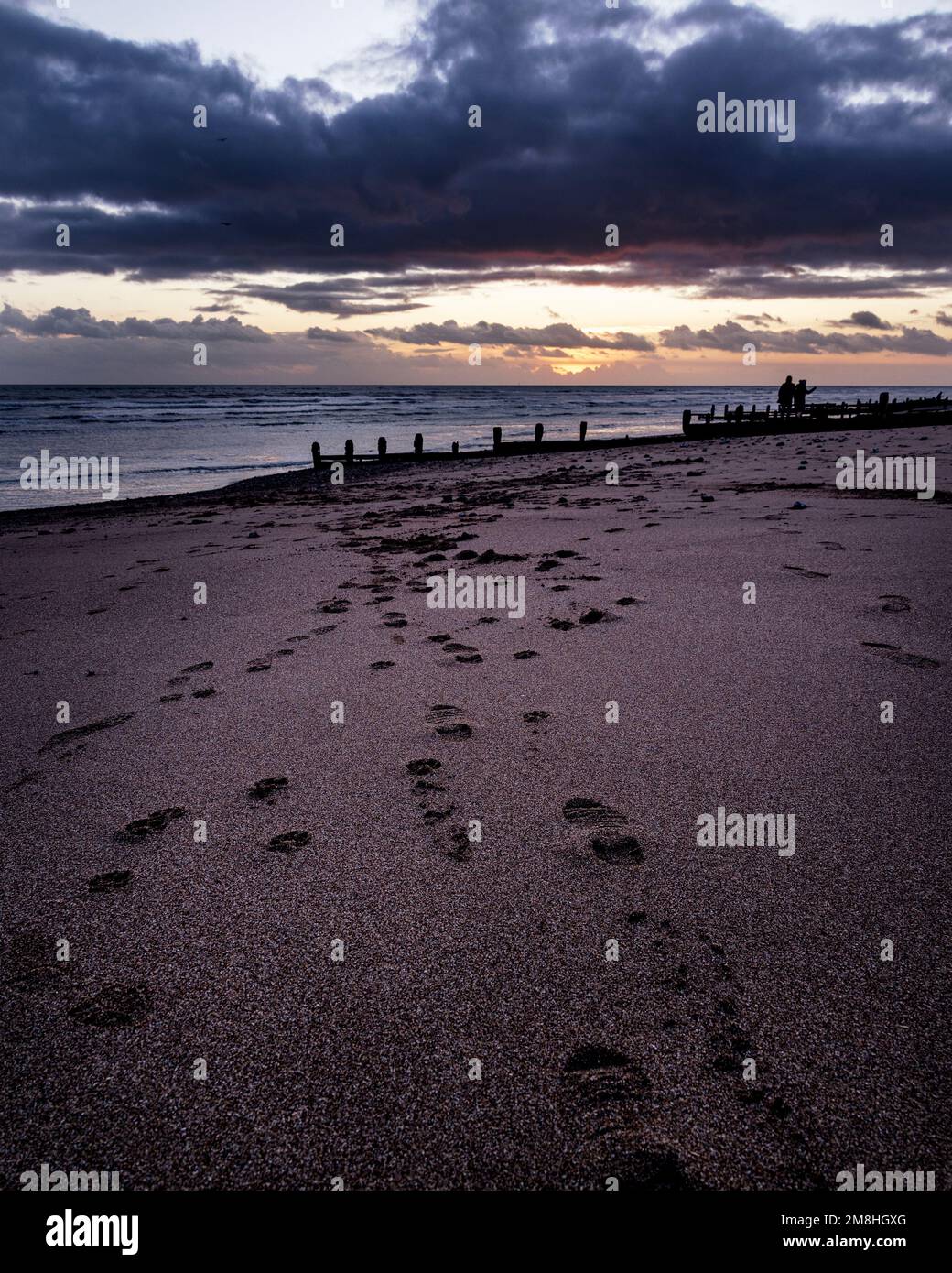 Footprints on the sand leading to the silhouette of two people at sunset on Worthing beach. Stock Photo