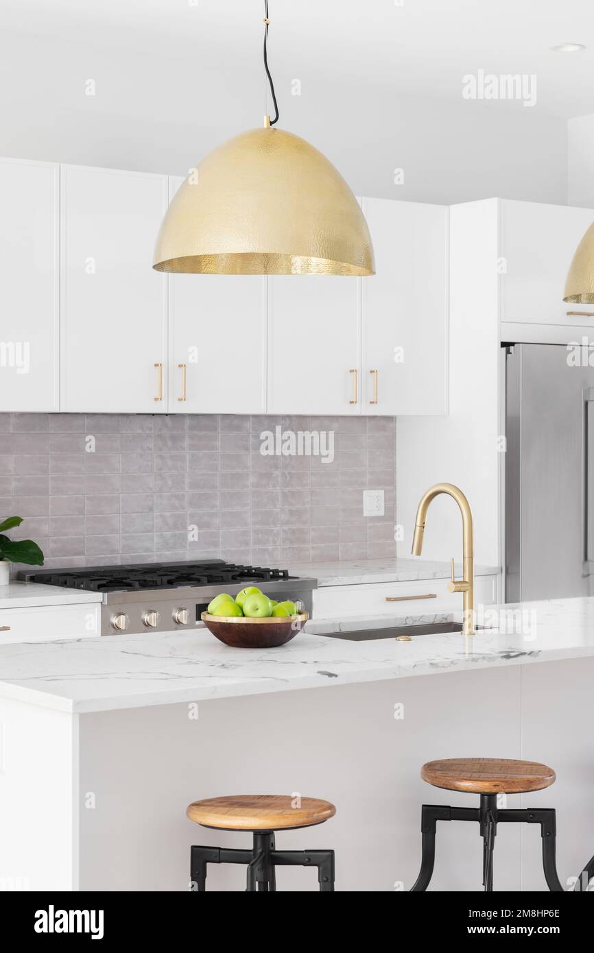 A kitchen detail with white cabinets, gold faucet and light hanging over the island with bar stools, and a tiled backsplash. Stock Photo