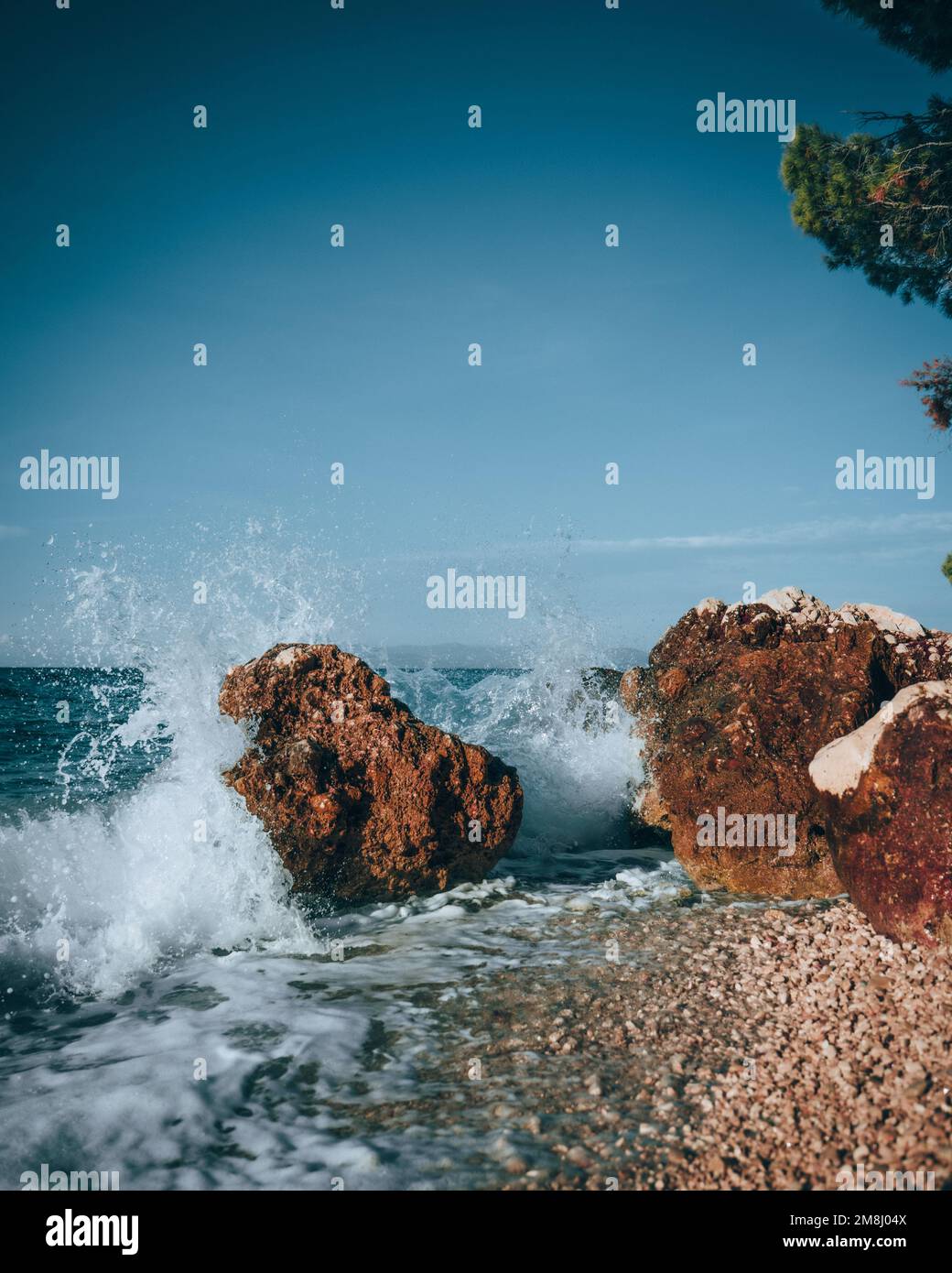 A vertical of water hitting stones on the beach, clear and sunlit sky