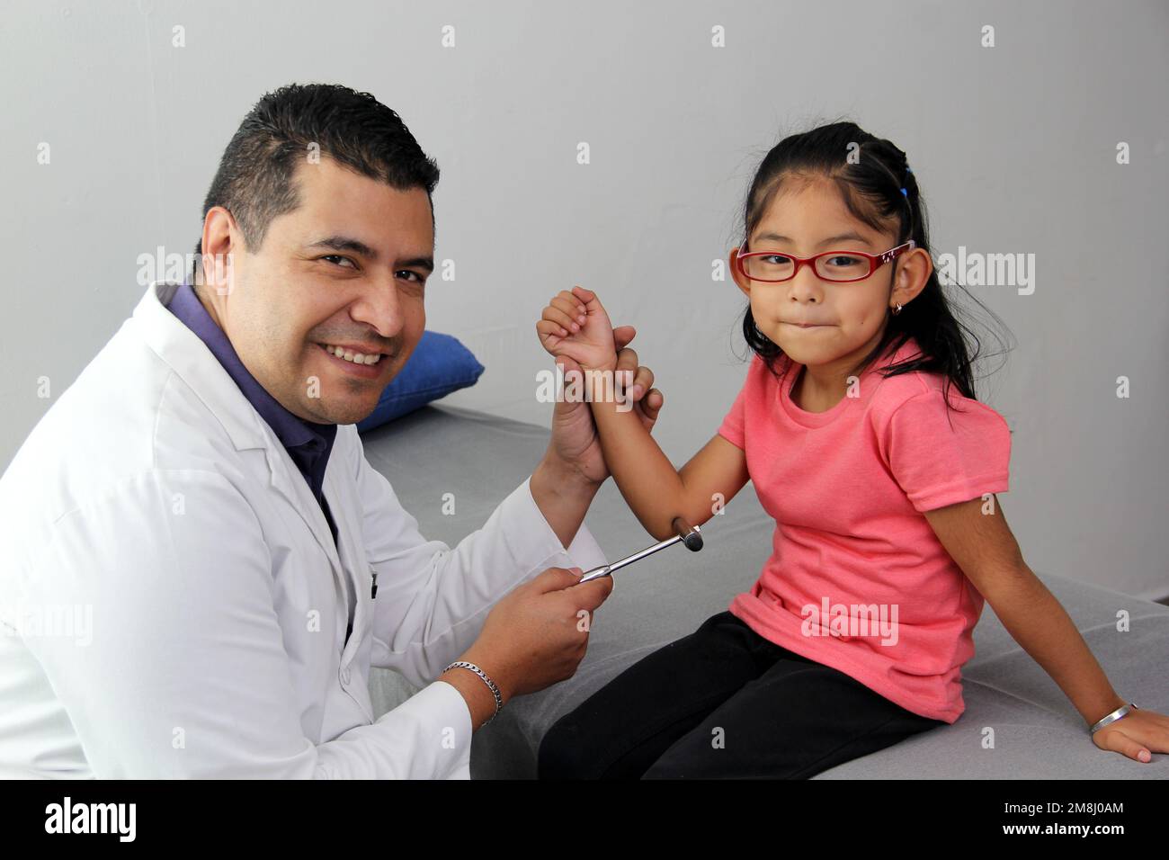 Latino doctor medic and girl patient in medical office checks her reflexes on hammer in her checkup to find disease diagnosis Stock Photo