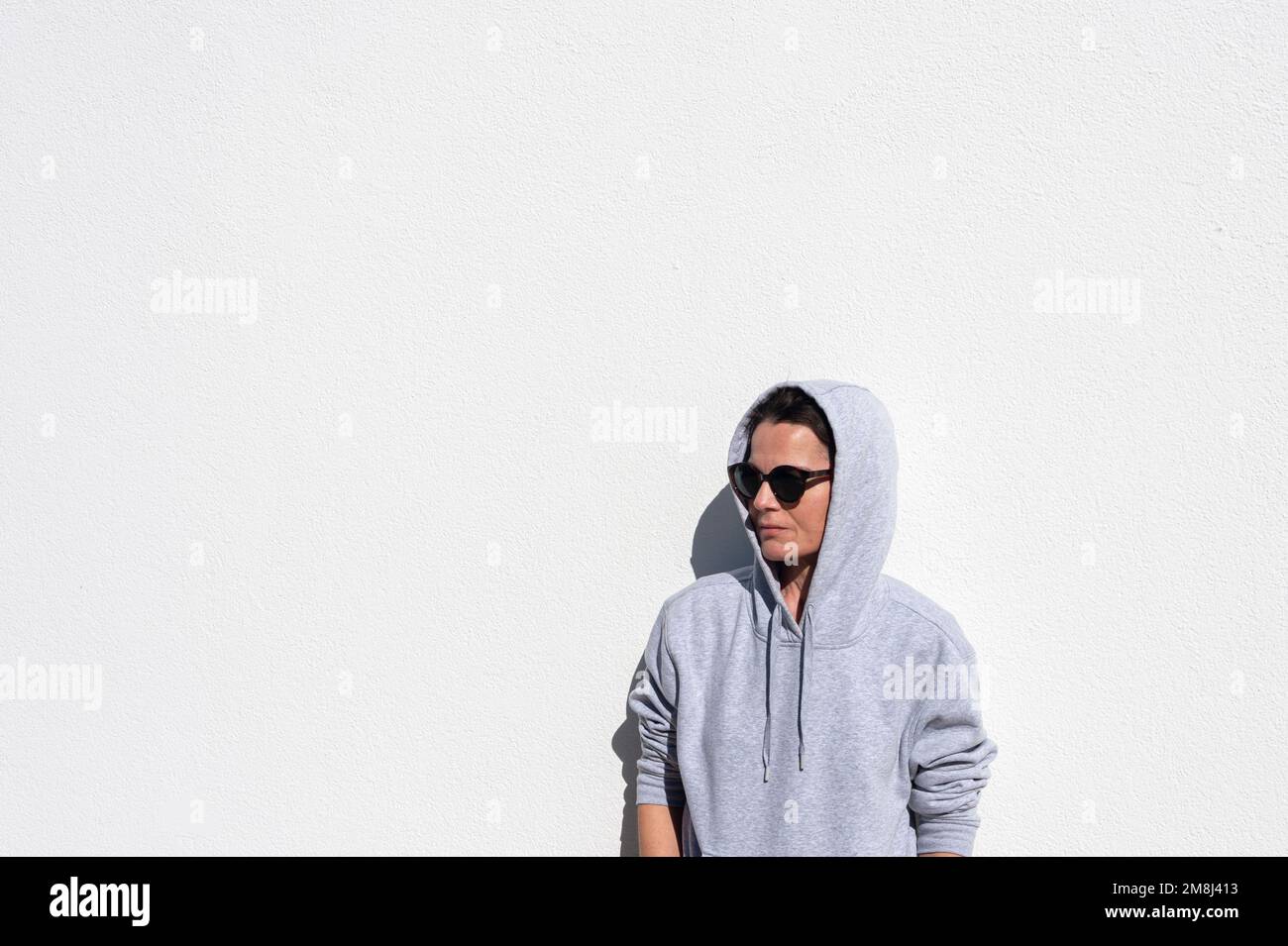 female wearing a grey hoodie leaning a gainst a white wall in the sunshine. Stock Photo