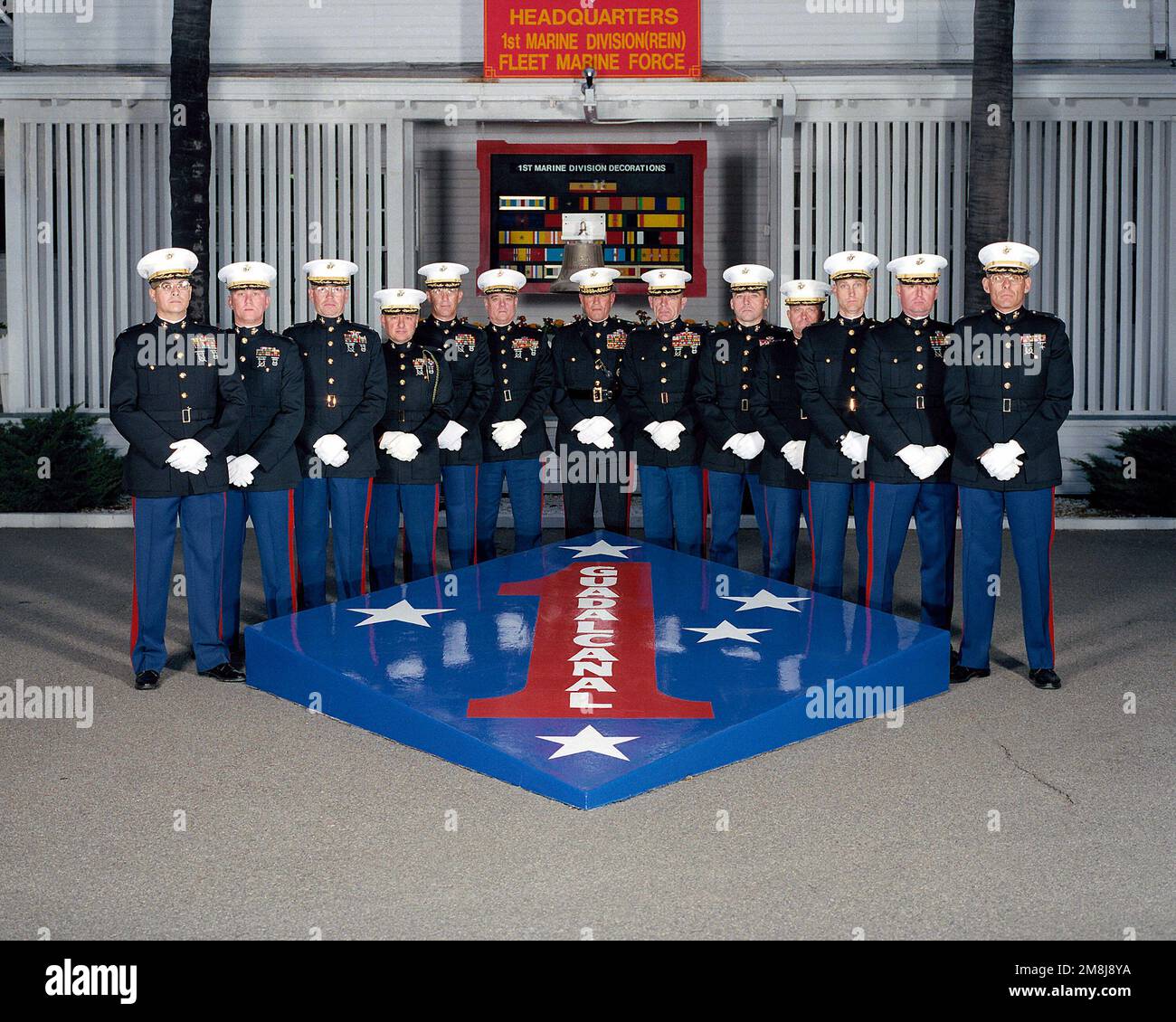 Exterior, MLS, MG Charles E. Wilhelm, USMC, Commanding General 1ST Marine Division and 12 Regimental Commanders, in dress blues, line up on two sides of a large replica of the 1ST Marine Division Patch and Seal. In background behind the staff is a sign, 'HEADQUARTERS 1ST MARINE DIVISION (REIN) FLEET MARINE FORCE'. Behind the staff is a large plaque showing the 1ST marine Division's Decorations and a ship's bell. The bell is from the USS WHARTON (AP-7) a WW II Troop Transport. The ship was named for the third Commandant of the Marine Corps, LTC Frank Wharton. Base: Marine Corps Base Camp Pendle Stock Photo