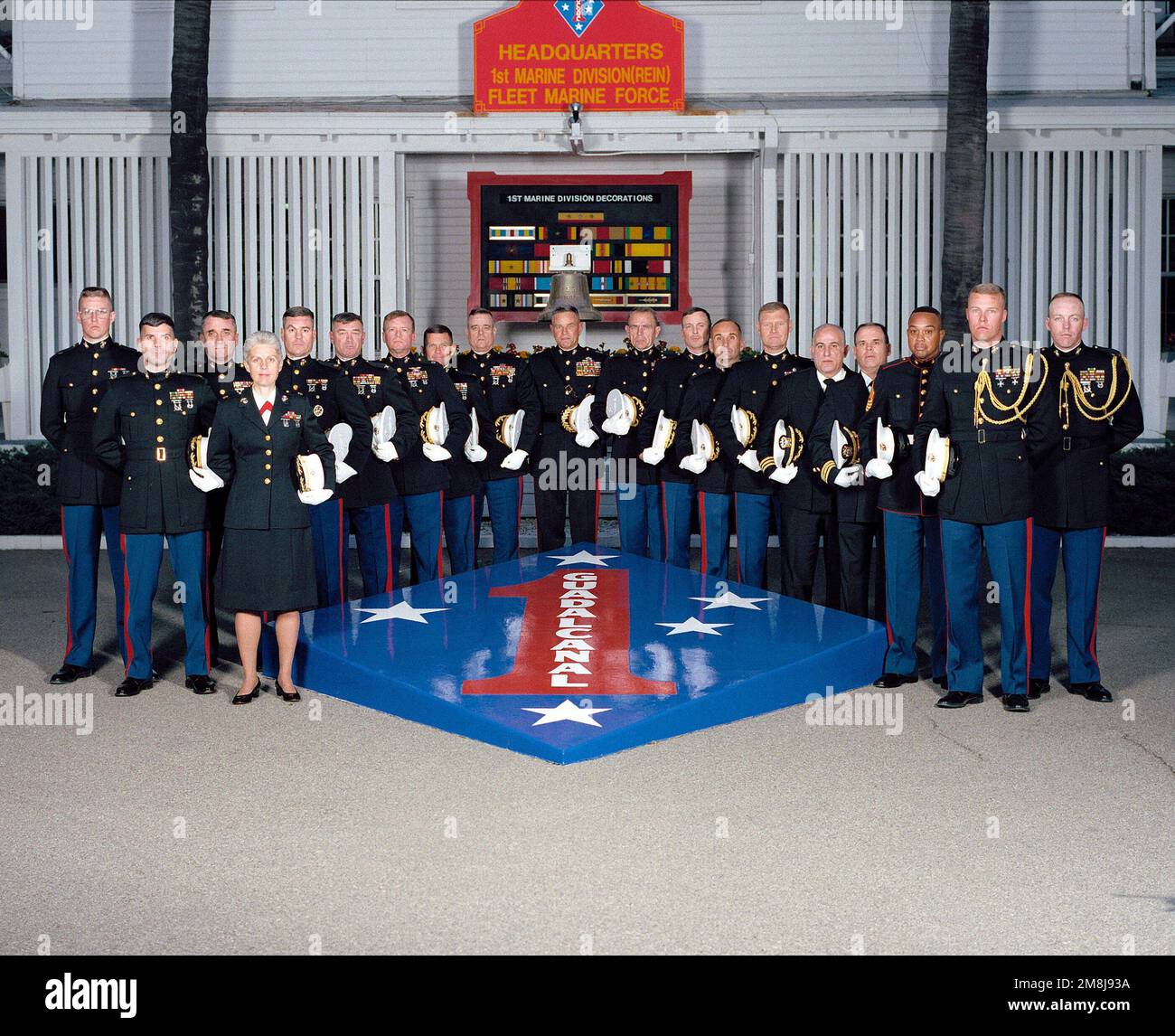 Exterior, MLS, MG Charles E. Wilhelm, USMC, Commanding General 1ST Marine Division and 18 members of his principal staff, in dress blues and holding hats, line up on two sides of a large replica of the 1ST Marine Division Patch and Seal. In background behind the staff is a sign, 'HEADQUARTERS 1ST MARINE DIVISION (REIN) FLEET MARINE FORCE'. Behind the staff is a large plaque showing the 1ST marine Division's Decorations and a ship's bell. The bell is from the USS WHARTON (AP-7) a WW II Troop Transport. The ship was named for the third Commandant of the Marine Corps, LTC Frank Wharton. Base: Mar Stock Photo