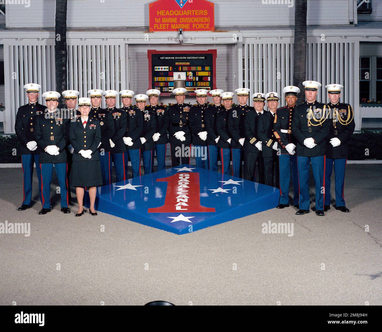 Exterior, MLS, MG Charles E. Wilhelm, USMC, Commanding General 1ST Marine Division and 18 members of his principal staff, in dress blues, line up on two sides of a large replica of the 1ST Marine Division Patch and Seal. In background behind the staff is a sign, 'HEADQUARTERS 1ST MARINE DIVISION (REIN) FLEET MARINE FORCE'. Behind the staff is a large plaque showing the 1ST marine Division's Decorations and a ship's bell. The bell is from the USS WHARTON (AP-7) a WW II Troop Transport. The ship was named for the third Commandant of the Marine Corps, LTC Frank Wharton. Base: Marine Corps Base Ca Stock Photo