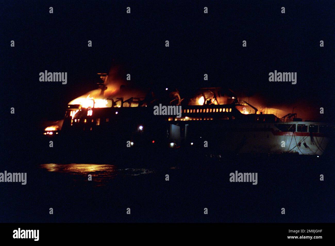 A port side view of the burning Egyptian ferry boat AL-QUAMAR AL-SAUDI AL-MISRI in the early morning hours before dawn as seen from the destroyer USS BRISCOE (DD-977). The ferry boat sent out a distress call after the ship's boiler had blown up and set the ship hopelessly on fire. The call to abandon ship was also given. The USS BRISCOE was first on the scene and moved in quickly to rescue survivors from the ship. Several survivors can be seen climbing down jacobs ladders at the ferry's fantail. Country: Northern Red Sea Stock Photo