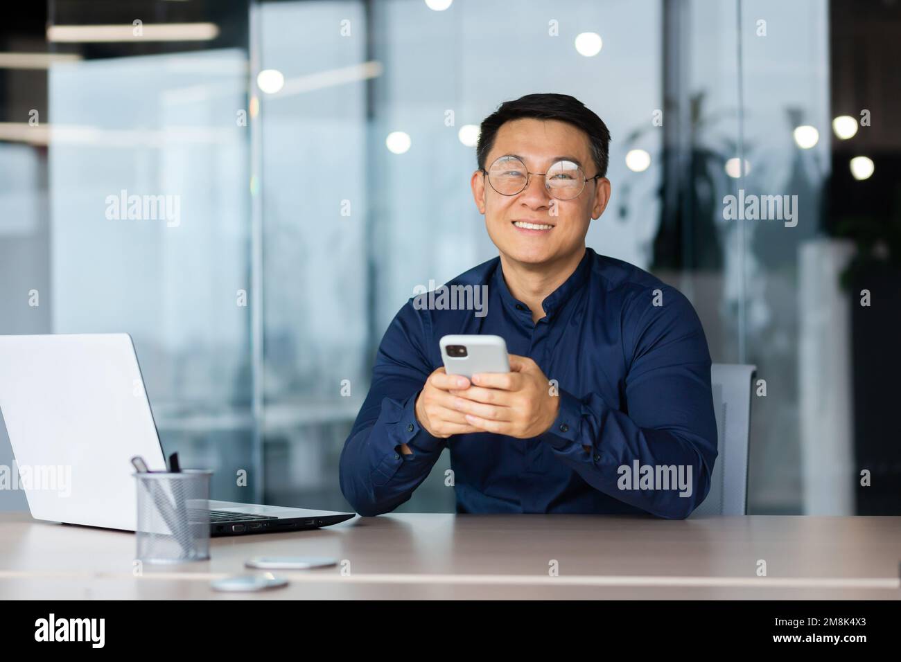 A young man in glasses sits in the office at a table with a laptop, holds a phone in his hands, smiles at the camera. Stock Photo