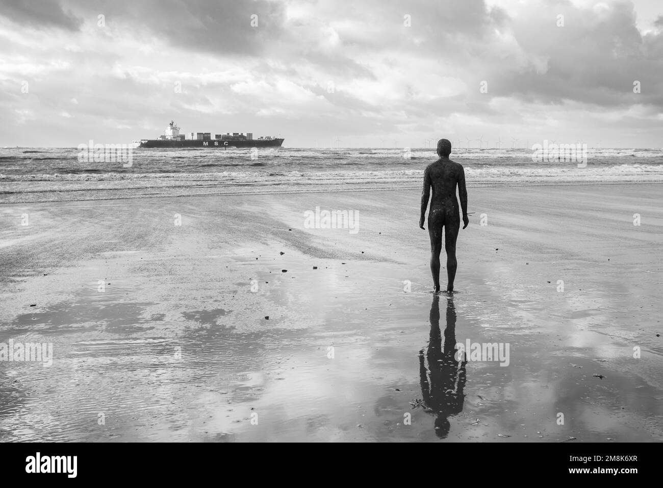 One of the Iron Men at Another Place on Crosby beach near Liverpool watches a large container ship exiting the River Mersey in January 2023. Stock Photo