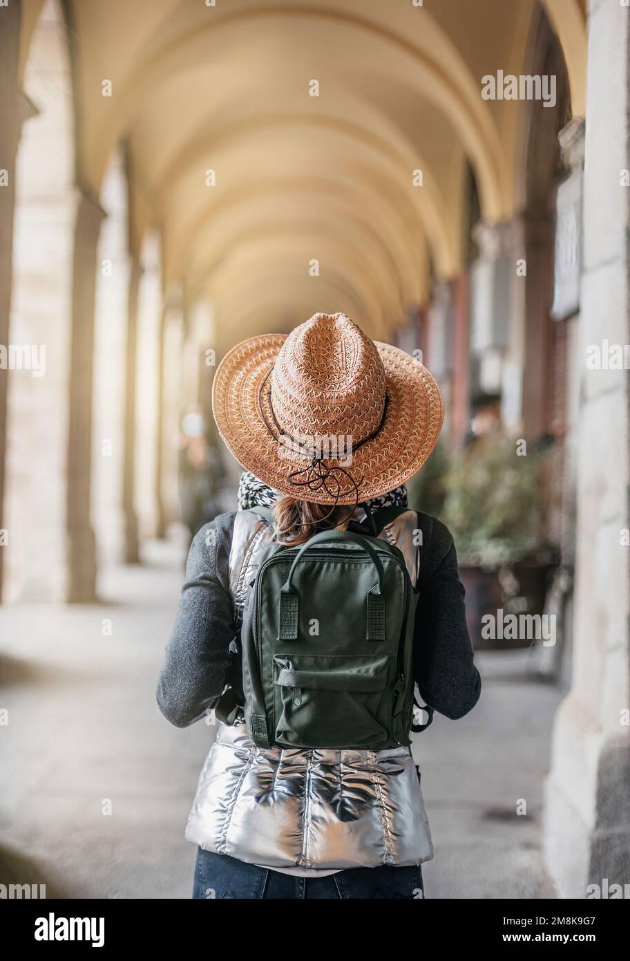 a woman hiker with her back turned, walks inside a long corridor of arches and columns in an old square in Madrid. she wears a straw hat and carries Stock Photo