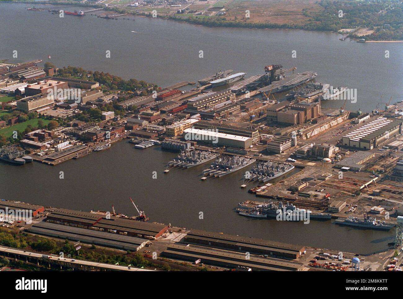 An aerial view of the Philadelphia Naval Shipyard looking southeast. The shipyard closed on September 30, 1995, but the Navy Intermediate Ship Maintenance Facility (NIMSF) will continue to store decommissioned and mothballed ships. Visible are two aircraft carriers, two amphibious assault ships, a heavy cruiser, five supply ships and numerous destroyers and frigates. Base: Philadelphia State: Pennsylvania (PA) Country: United States Of America (USA) Stock Photo