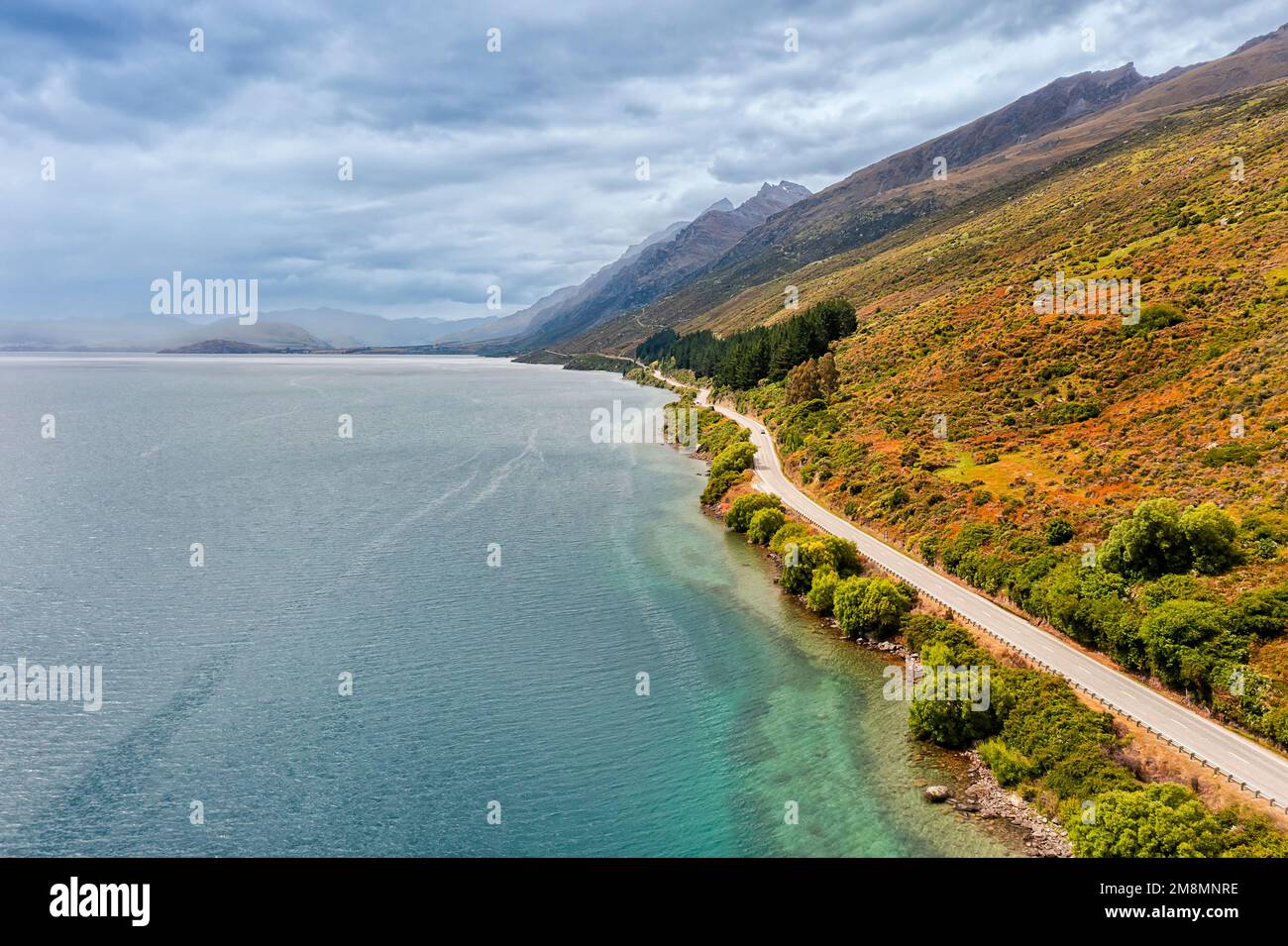 Remote highway along Wakatipu lake in South island of New Zealand under scenic mountain ranges on a cloudy sunny day. Stock Photo