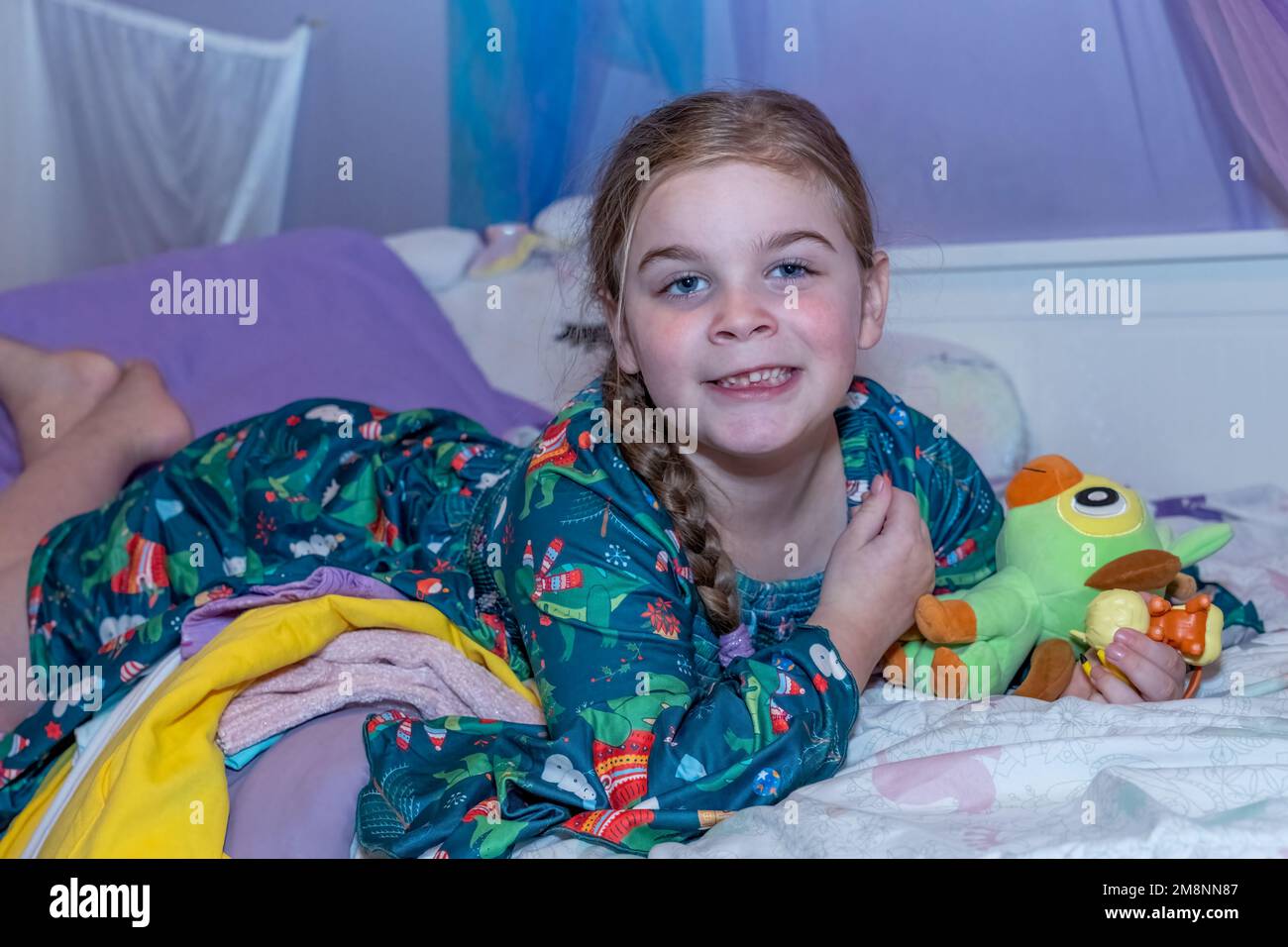 Smiling six year old girl missing front teeth lying on her bed with her stuffed animals. Stock Photo