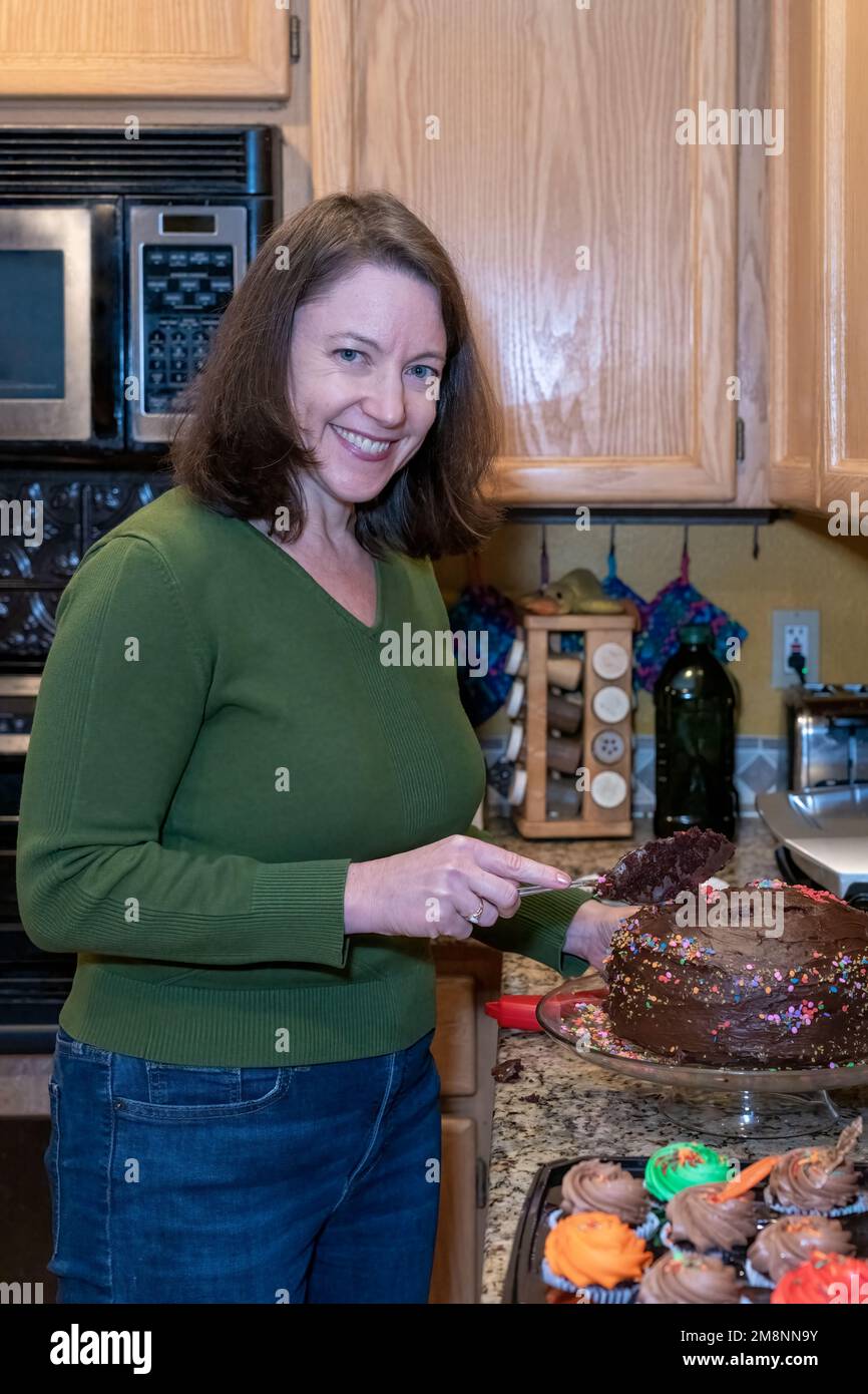 Mother cutting a chocolate birthday cake in her kitchen, with cupcakes in the foreground. (MR) Stock Photo