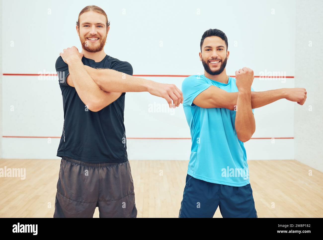 Portrait of two squash players stretching and smiling before playing court game. Happy fit caucasian and mixed race athlete standing together and Stock Photo