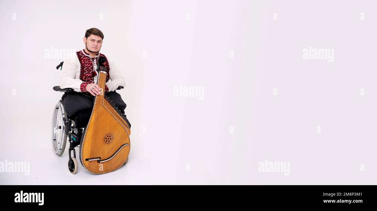 invalid handsome young man with kobza musical instrument in hands sits on white background on wheelchair without legs war veteran dressed in Ukrainian Stock Photo