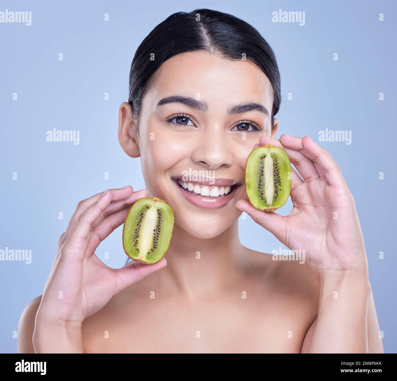 Studio Portrait of a happy smiling mixed race woman holding a kiwi fruit. Hispanic model promoting the skin benefits of a healthy diet against a blue Stock Photo