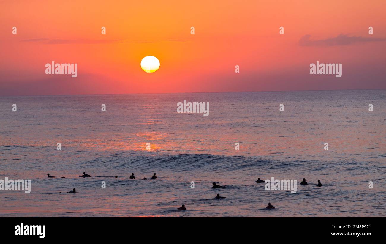 Ocean horizon sea waters with sun rising in the sky orange red colors with blurred surfers silhouetted surfing morning waves. Stock Photo
