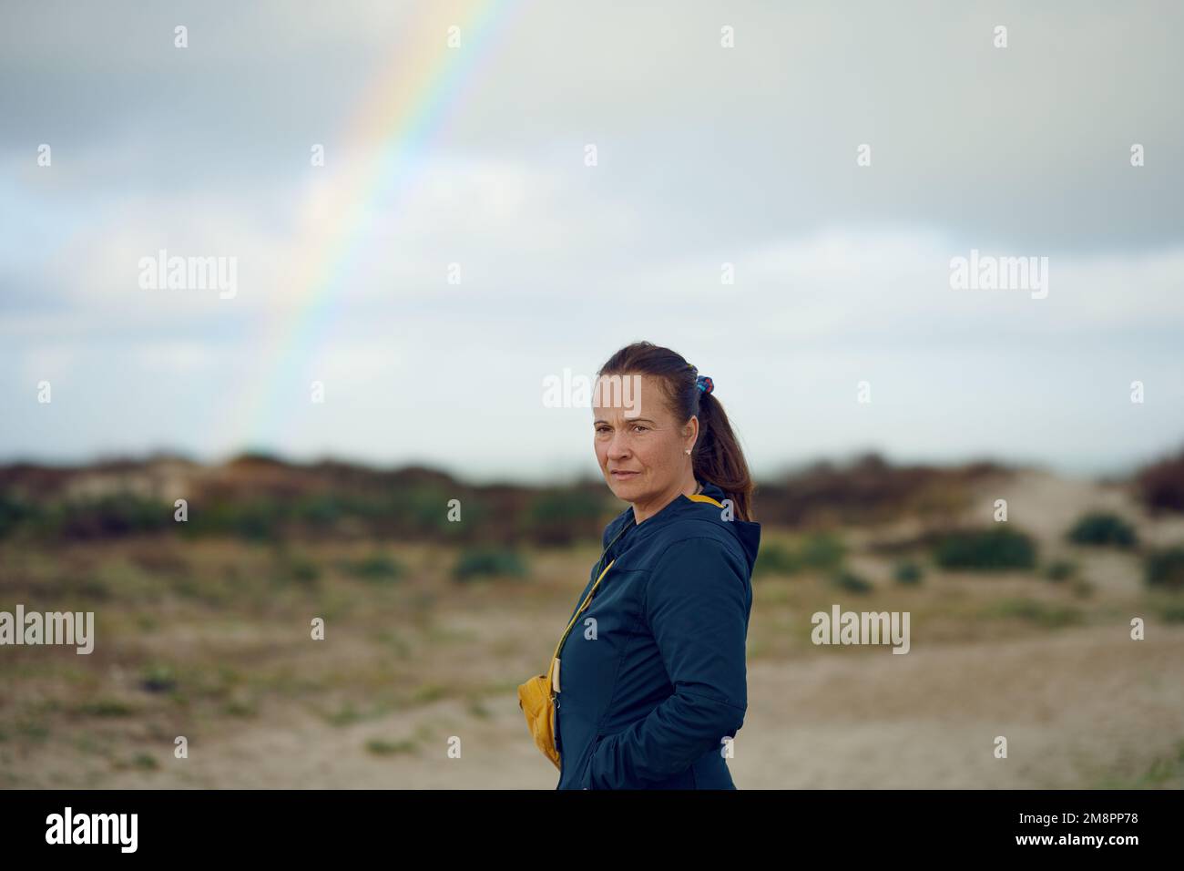Middle-aged woman looking thoughtful at the camera with a rainbow in the background, concept for future outlook Stock Photo