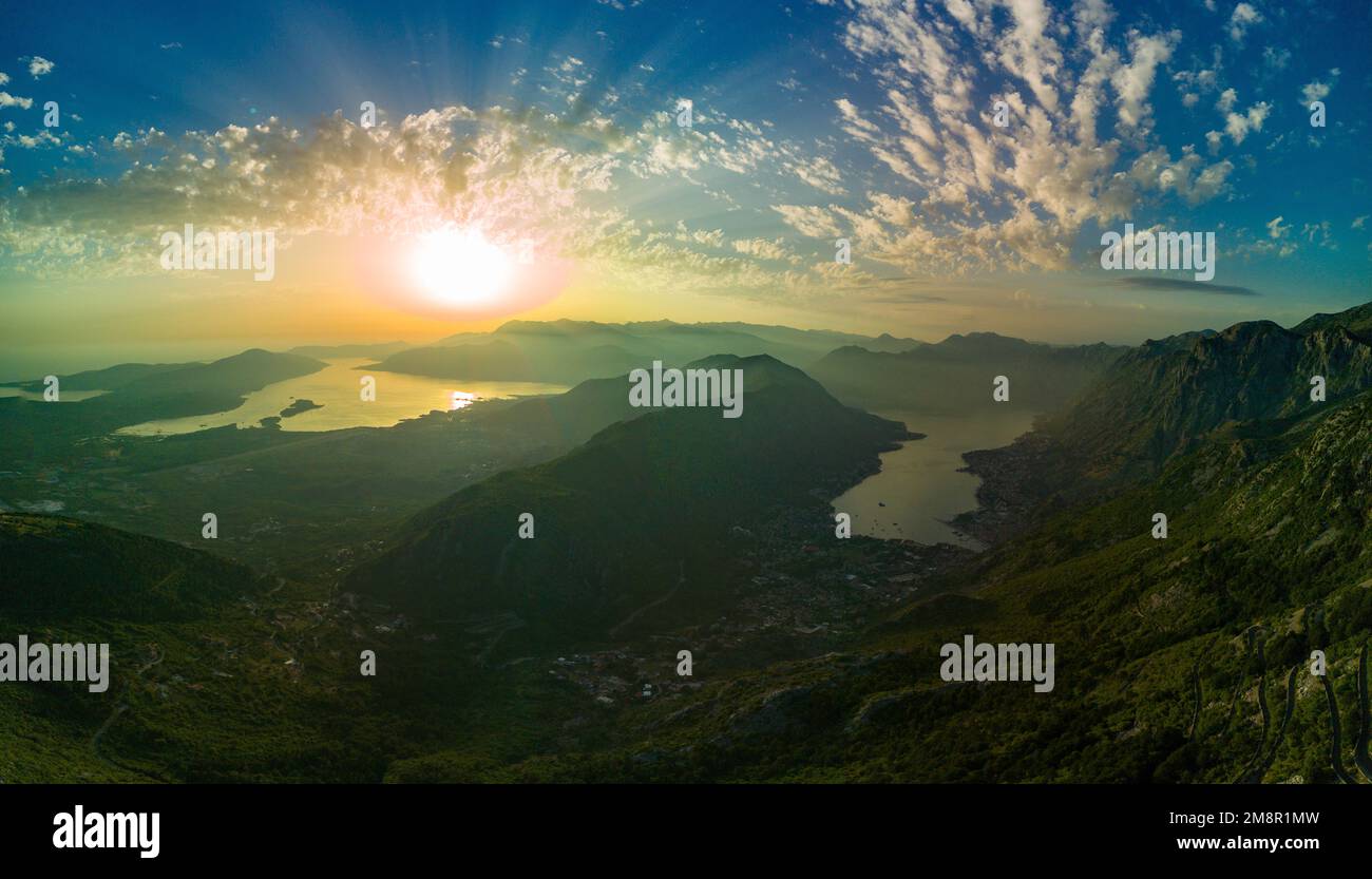 Panorama of bright dazzling golden sun in the blue cloudy evening sky illuminates all the silhouettes of the peaks of the Balkan Montenegrin mountains Stock Photo