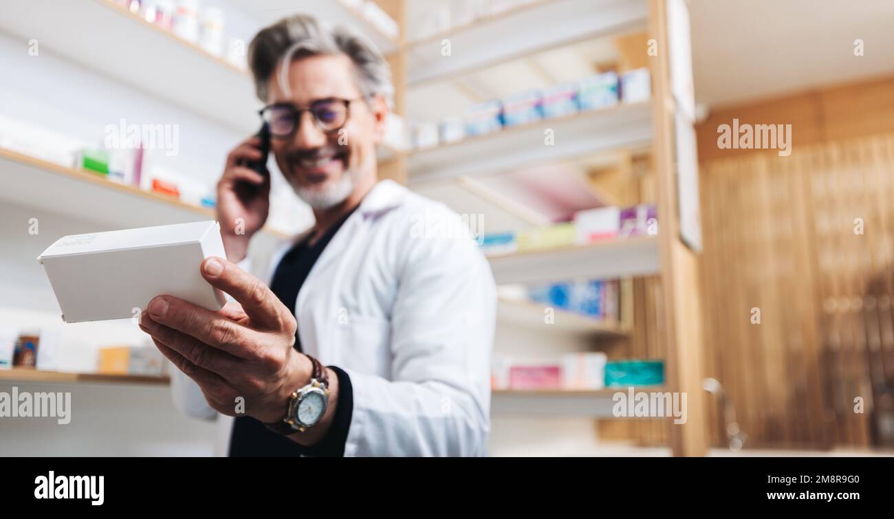 Drug store pharmacist holding a box of pills and talking to a patient on the phone. Senior healthcare worker reading medication label to confirm a pre Stock Photo