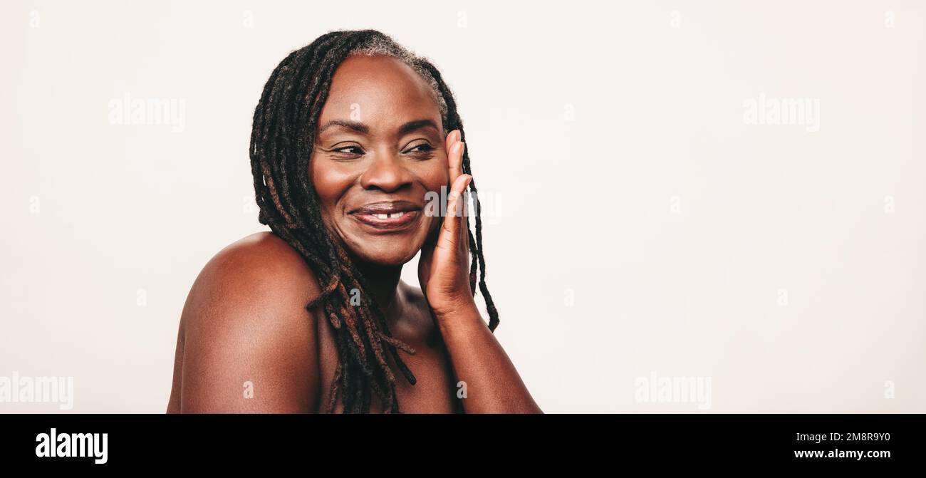 Happy mature woman with dreadlocks smiling while touching her flawless skin. Cheerful dark-skinned woman embracing her beautiful melanated skin. Matur Stock Photo