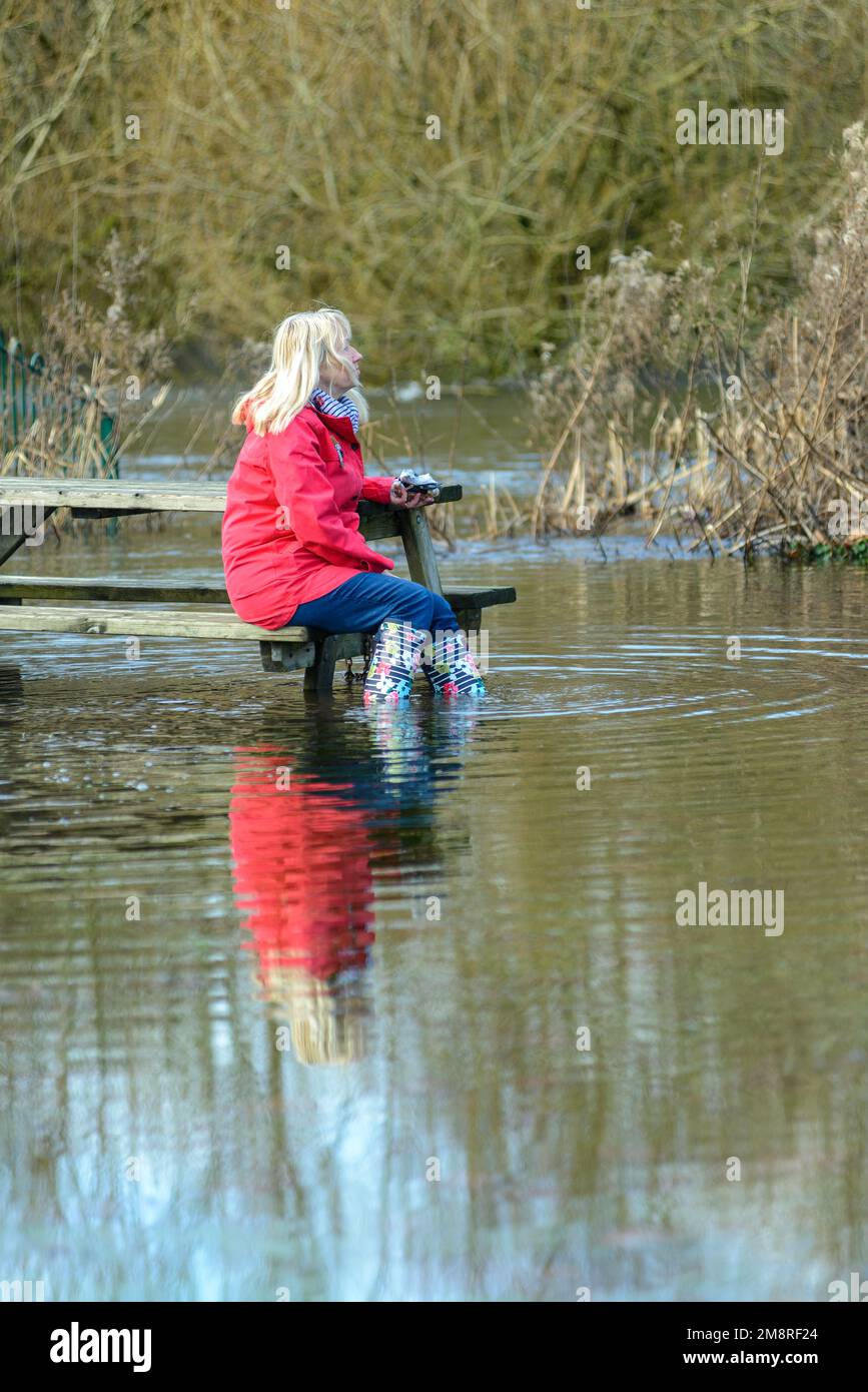 Woman sitting at flooded park bench with Wellington boots in water Stock Photo