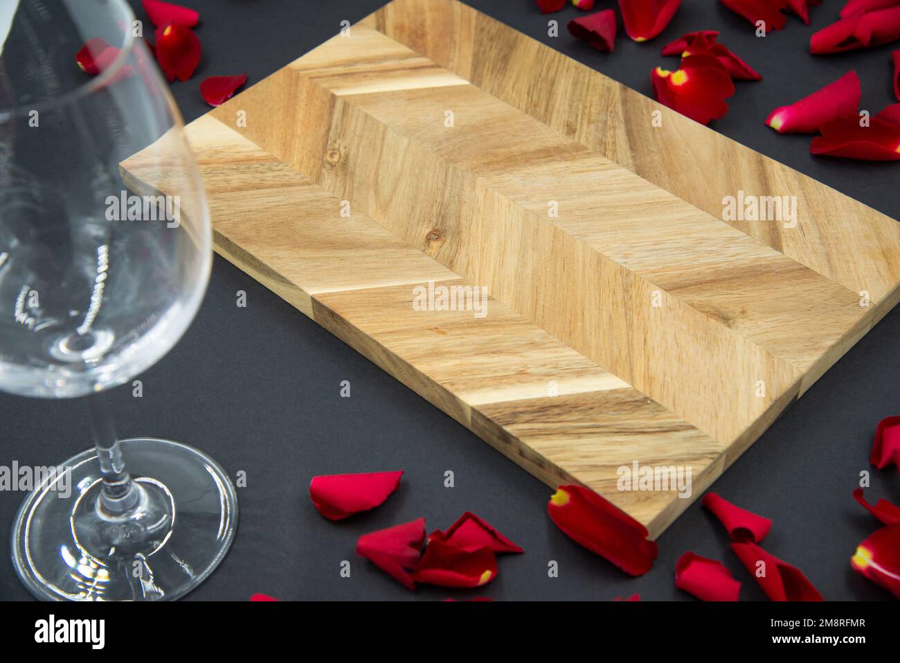 Wooden cutting board for cooking on a dark black background with a glass of wine and red rose petals. Stock Photo