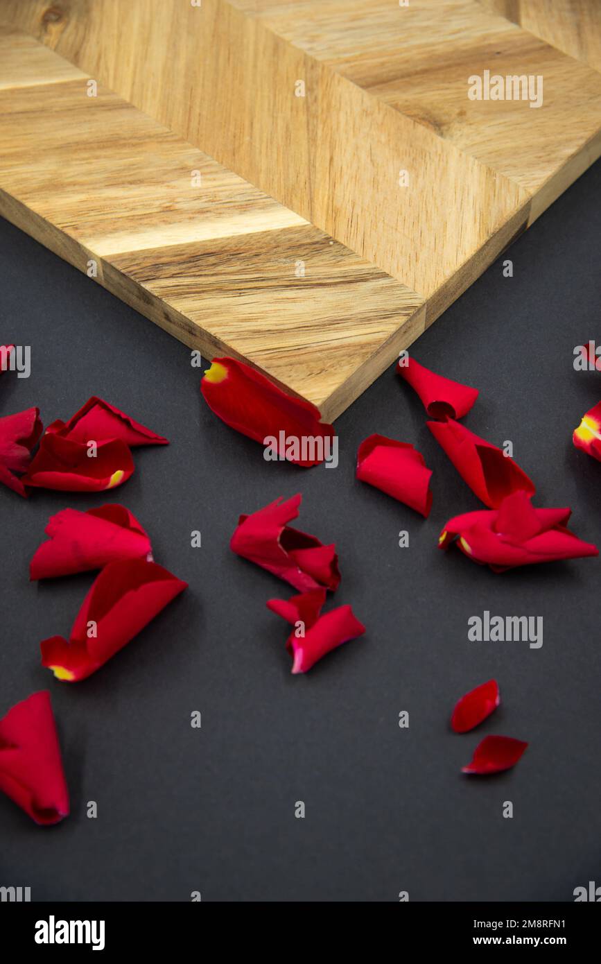 Red rose petals on a black background next to a wooden board for cooking. Stock Photo