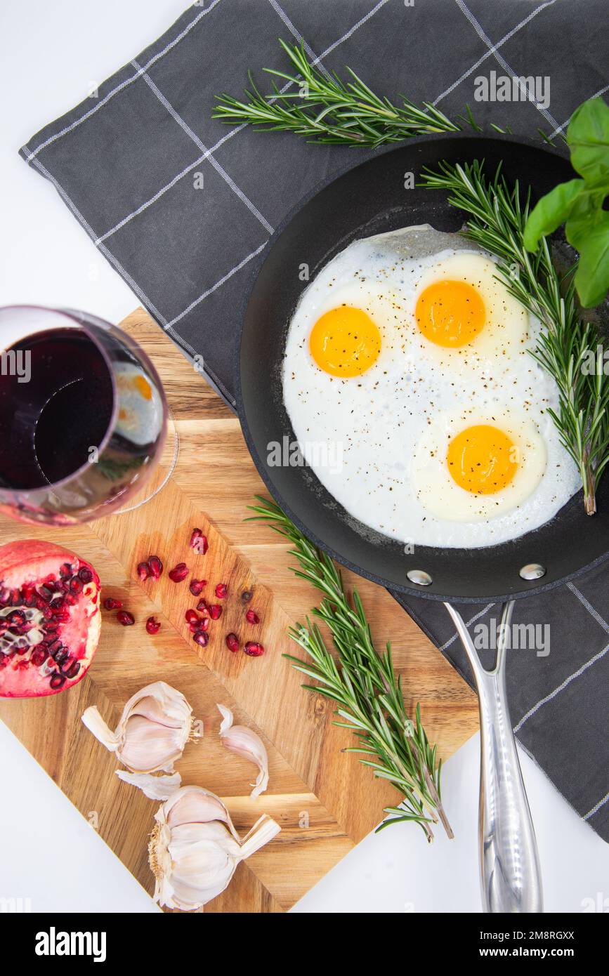 breakfast or brunch fried eggs in a black pan and red wine in a crystal glass and red pomegranate with garlic and basil. view from the top. Stock Photo
