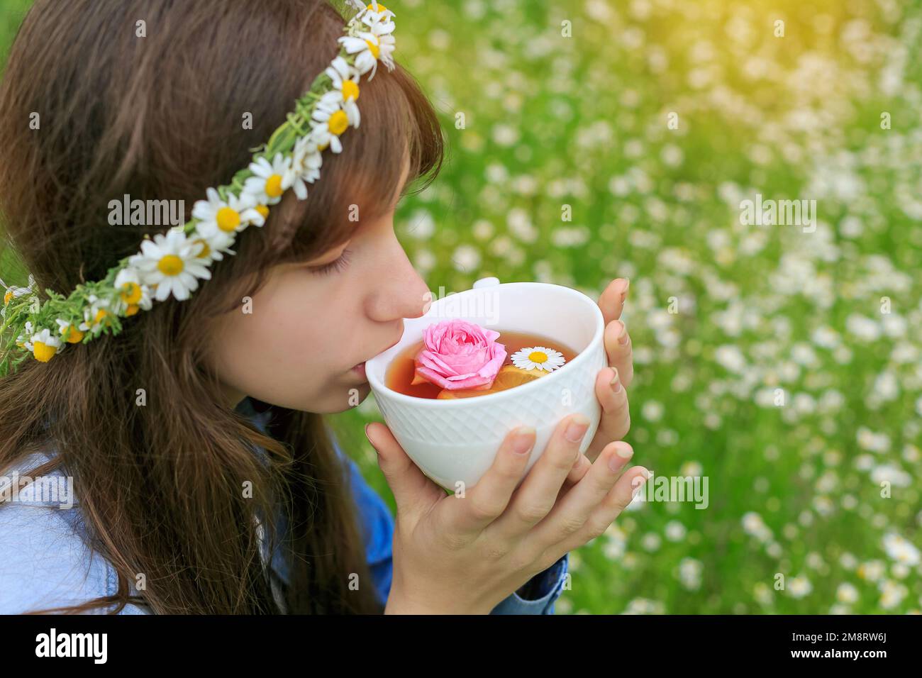 Brunette healthy woman drinking fragrant herbal rose tea. Selective focus. Teenager girl in flower wreath holding white cup with chamomile detox drink Stock Photo