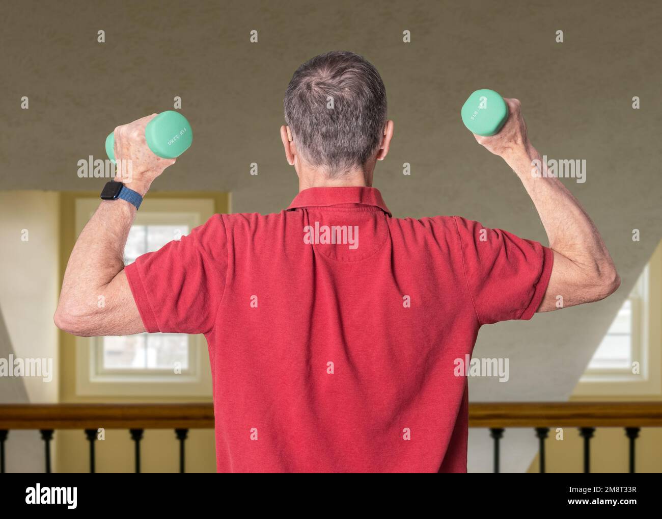 Senior man in red shirt seen from back and exercising with dumbbells in home against railing Stock Photo