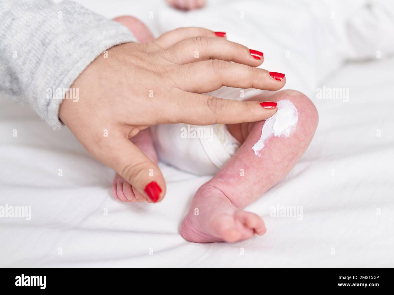 Adorable caucasian baby lying on bed applying lotion on leg at bedroom Stock Photo